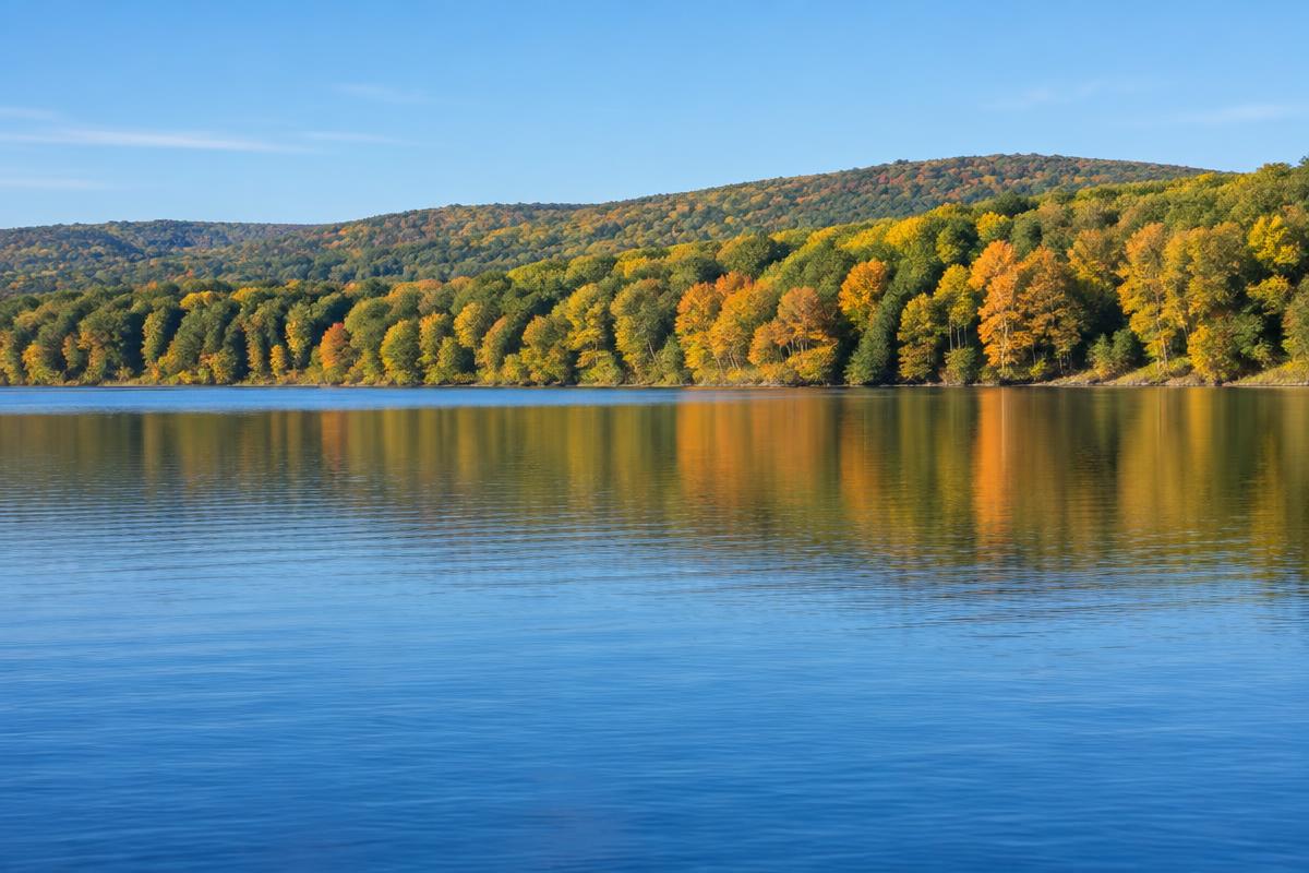 Calm blue Pocono lake beneath an early-autumn hardwood shoreline in soft morning light